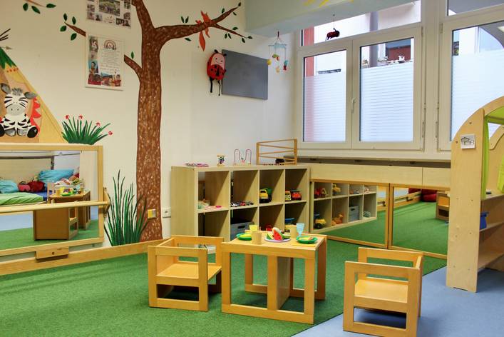 Ein Gruppenraum im Krippenbereich des Kifaz. Im Zentrum steht eine Kindersitzgruppe und im Hintergrund ein Regal und an der Wand ist ein großer gemalter Baum 